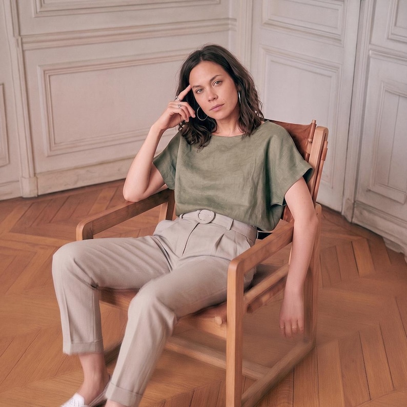 May include: A sage green short-sleeved top with a wide neckline and a pair of light beige high-waisted trousers featuring a matching fabric belt with a round buckle, a pleated front, and cuffed hems, worn while seated in a wooden chair.