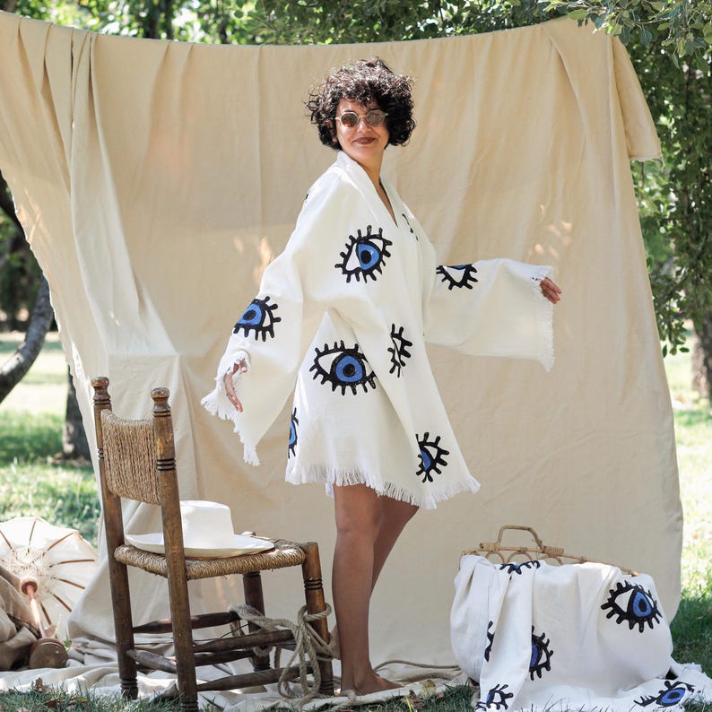 Op de afbeelding: Een witte kimono-stijl badjas met een herhalend patroon van blauwe en zwarte oogmotieven. Het kledingstuk heeft wijde, losse mouwen en een zoom afgewerkt met franjes. De stof is gebroken wit met een textuur over het gehele oppervlak.