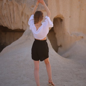 Puede incluir: Una blusa blanca de manga corta con tejido texturizado y escote profundo en la espalda, combinada con pantalones cortos negros de talle alto con cintura el&aacute;stica y dobladillo con volantes, junto a sandalias de tiras color canela.