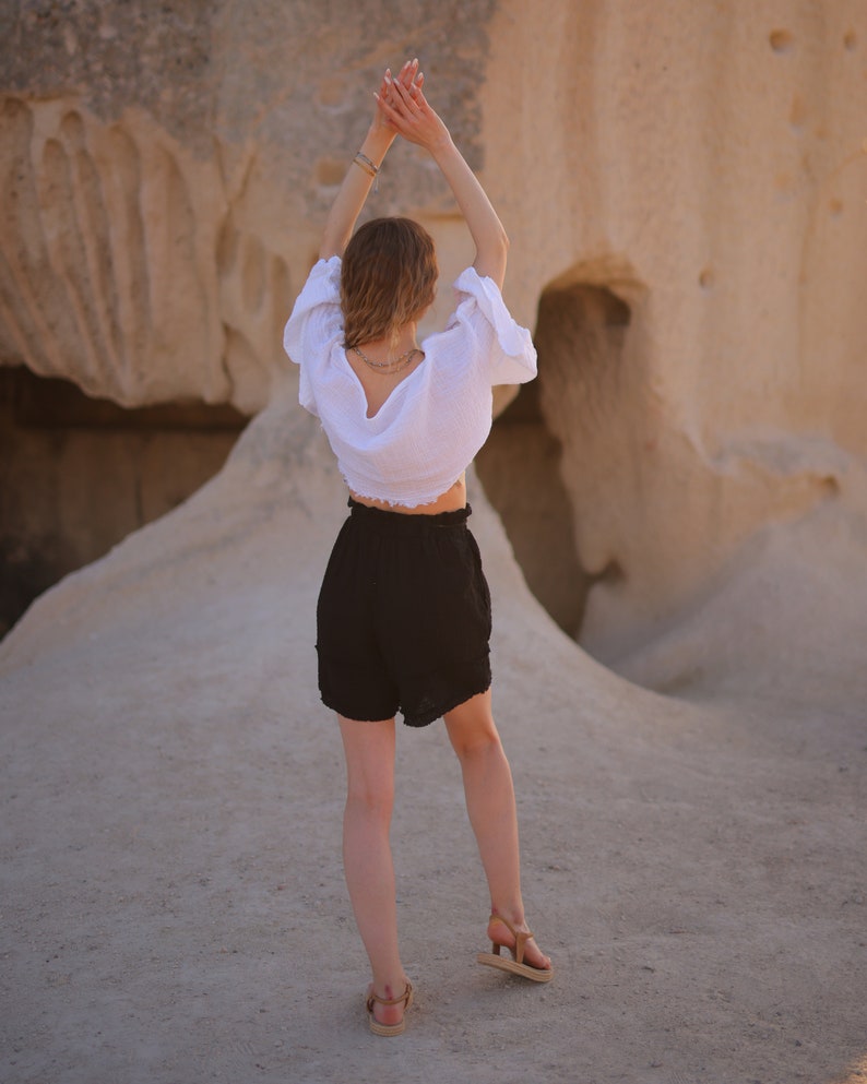 Puede incluir: Una blusa blanca de manga corta con tejido texturizado y escote profundo en la espalda, combinada con pantalones cortos negros de talle alto con cintura el&aacute;stica y dobladillo con volantes, junto a sandalias de tiras color canela.