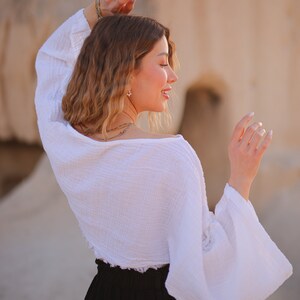 May include: A woman wearing a white, loose-fitting, cropped top with long sleeves and a black skirt. The top has a ruffled hem and the sleeves are gathered at the wrists.