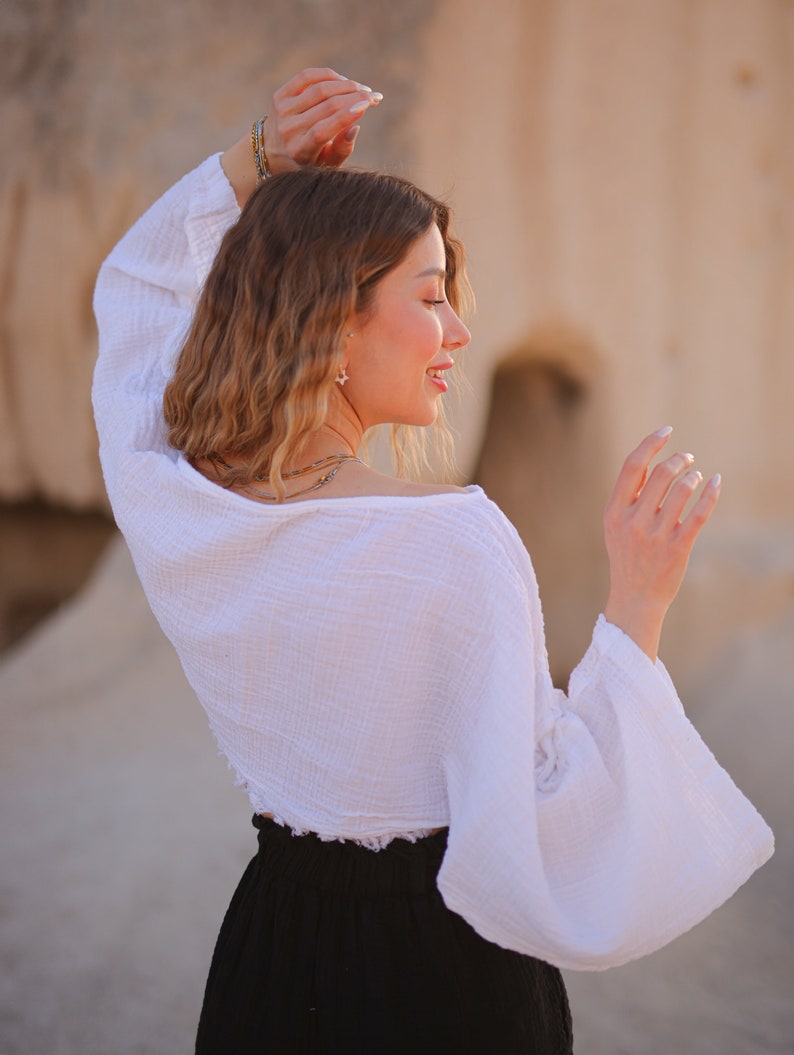 May include: A woman wearing a white, loose-fitting, cropped top with long sleeves and a black skirt. The top has a ruffled hem and the sleeves are gathered at the wrists.