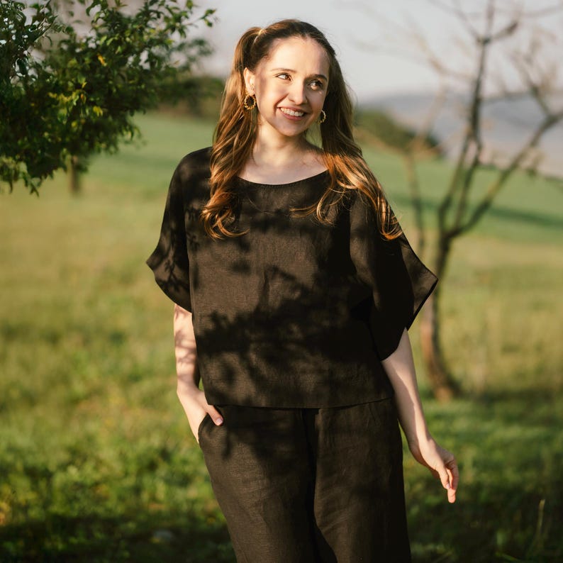 May include: A black short-sleeved top with a wide, rounded neckline and matching black trousers. The top features loose, flowing sleeves and a straight hemline, while the trousers have a relaxed fit with visible side pockets.