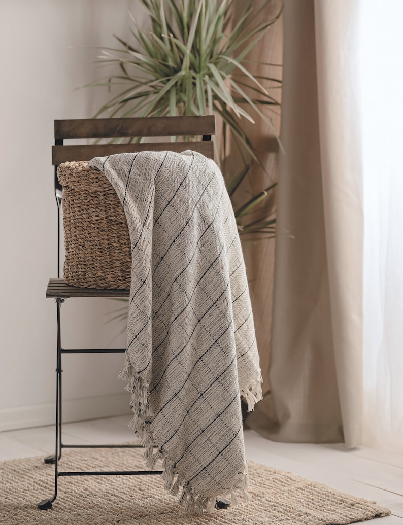Puede incluir: Una cesta tejida se encuentra sobre una silla de metal negro con una manta a rayas blancas y grises drapeada sobre el respaldo. La silla est&aacute; sobre una alfombra tejida frente a una pared blanca.