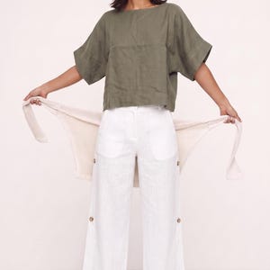 May include: An olive green short-sleeved top with a wide neckline paired with white wide-leg trousers featuring side button details and a cream-colored knit cardigan draped across the back, creating a layered ensemble with a clean aesthetic.