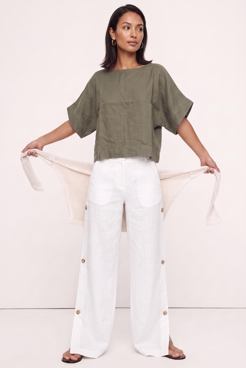 May include: An olive green short-sleeved top with a wide neckline paired with white wide-leg trousers featuring side button details and a cream-colored knit cardigan draped across the back, creating a layered ensemble with a clean aesthetic.