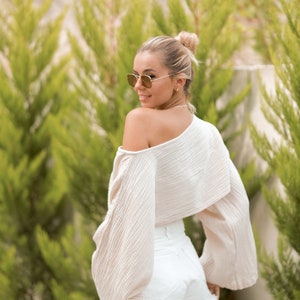 May include: A woman wearing a white off-the-shoulder top with long sleeves and white denim shorts. She is wearing sunglasses and has her hair pulled back in a bun.