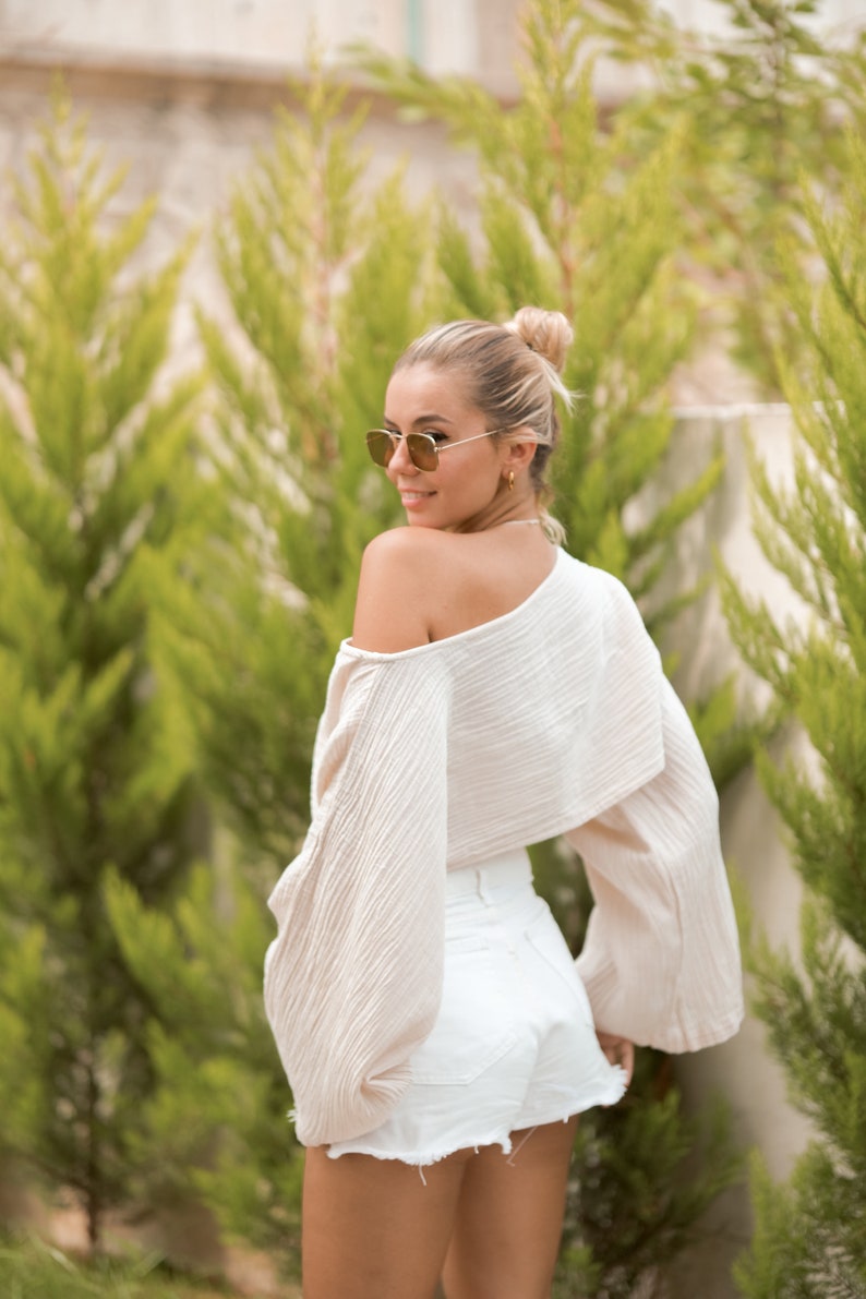 May include: A woman wearing a white off-the-shoulder top with long sleeves and white denim shorts. She is wearing sunglasses and has her hair pulled back in a bun.