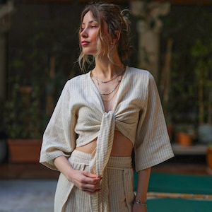May include: A woman wearing a beige and white striped crop top with a knotted front and matching pants. She is wearing multiple gold necklaces.