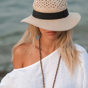 May include: A beige straw fedora hat with a black ribbon band, paired with a long necklace featuring small brown wooden beads and a white off-the-shoulder top. The hat has a woven texture and a wide brim that partially obscures the upper face.
