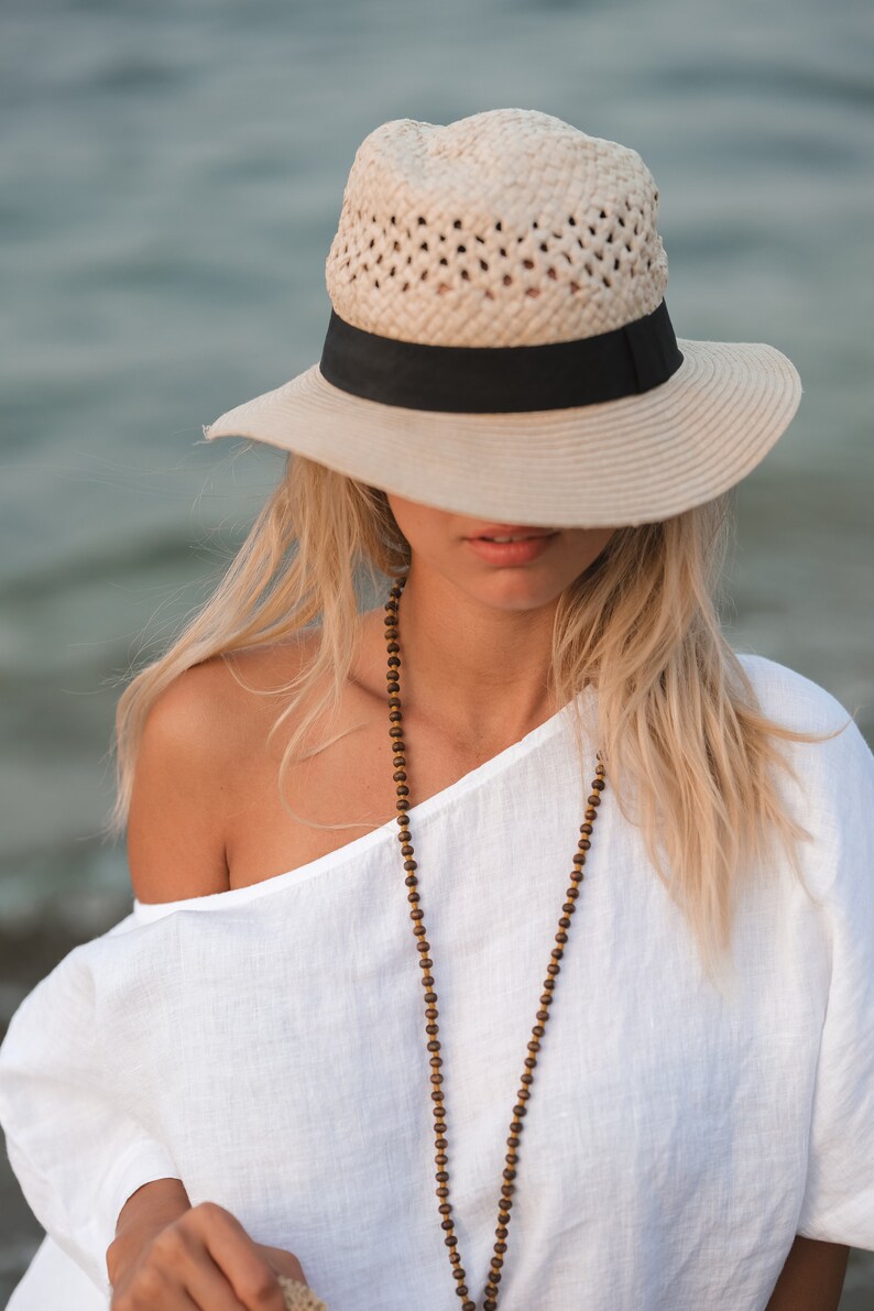 May include: A beige straw fedora hat with a black ribbon band, paired with a long necklace featuring small brown wooden beads and a white off-the-shoulder top. The hat has a woven texture and a wide brim that partially obscures the upper face.
