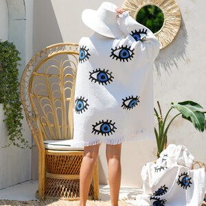 Op de afbeelding: Een wit oversized omslagdoek of handdoek met een herhalend patroon van blauwe en zwarte oogmotieven. Het kledingstuk heeft lange mouwen, een losvallend silhouet en een gefranjerde zoom. Een bijpassende witte hoed met brede rand wordt gedragen.