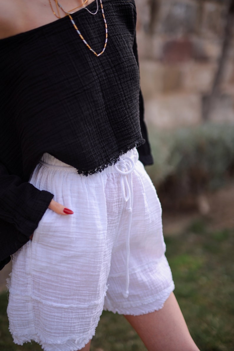 May include: White cotton shorts with a drawstring waist and frayed edges. The shorts are worn with a black off-the-shoulder top.