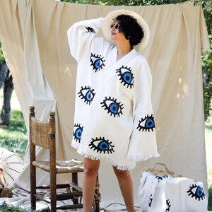 Op de afbeelding: Een gebroken witte kimono-stijl badjas met een herhalend patroon van geborduurde blauwe en zwarte ogen. Het kledingstuk heeft lange, wijde mouwen, een V-hals en een zoom afgewerkt met franjes. De stof is een effen, lichtgekleurd textiel.
