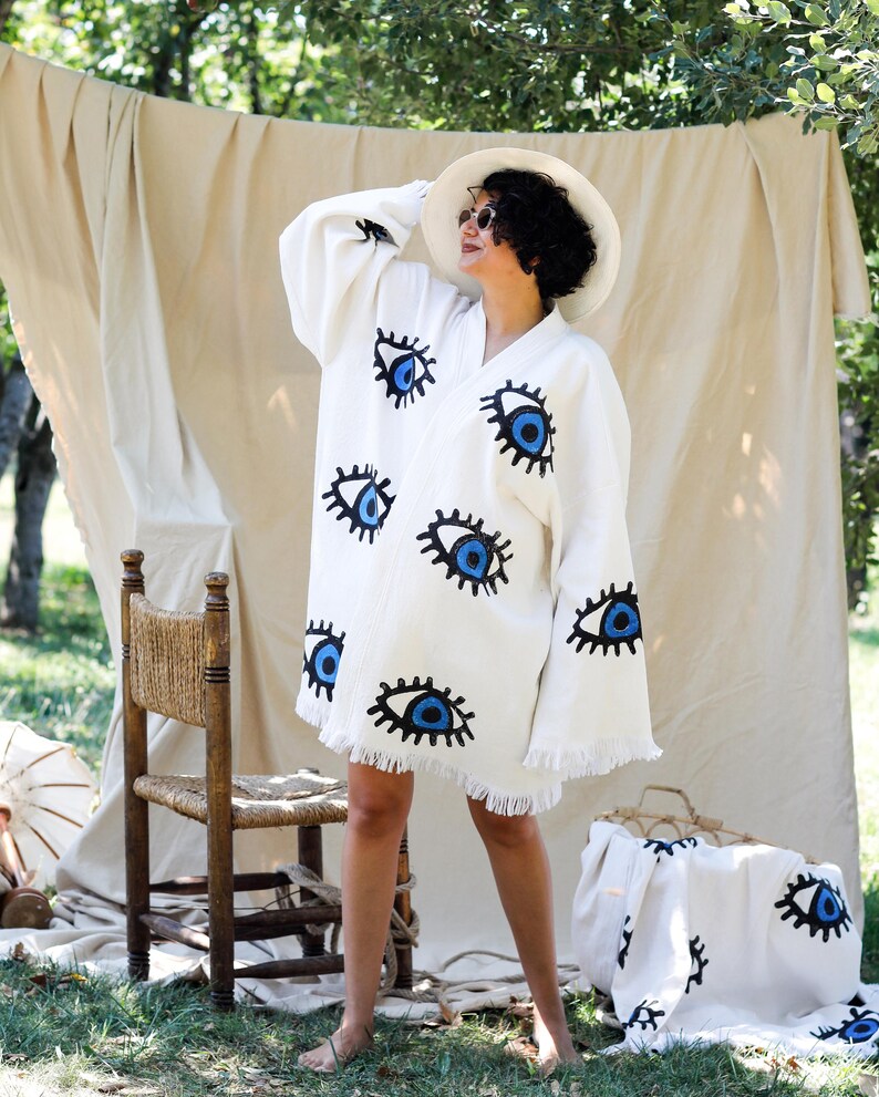 Op de afbeelding: Een gebroken witte kimono-stijl badjas met een herhalend patroon van geborduurde blauwe en zwarte ogen. Het kledingstuk heeft lange, wijde mouwen, een V-hals en een zoom afgewerkt met franjes. De stof is een effen, lichtgekleurd textiel.