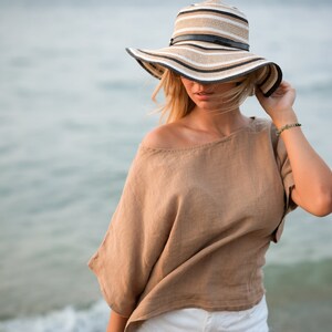 May include: A wide-brimmed sun hat featuring horizontal stripes in shades of tan, white, and black, accented with a thin black band around the crown. The hat is paired with a loose-fitting, short-sleeved tan top and white bottoms.