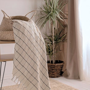 Puede incluir: Una manta de tiro blanca y negra a cuadros con flecos en los bordes, drapeada sobre una cesta de mimbre. La manta est&aacute; sobre una mesa de madera con una pata de metal negro. Una planta en maceta est&aacute; en el fondo.
