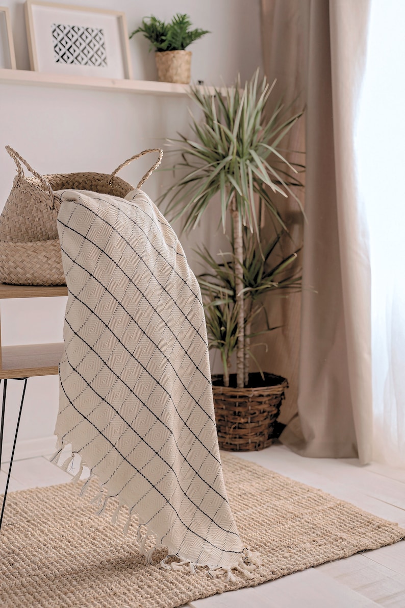 Puede incluir: Una manta de tiro blanca y negra a cuadros con flecos en los bordes, drapeada sobre una cesta de mimbre. La manta est&aacute; sobre una mesa de madera con una pata de metal negro. Una planta en maceta est&aacute; en el fondo.