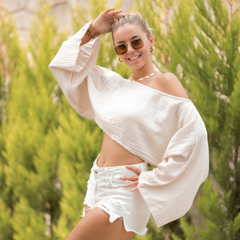 May include: A woman wearing a white off-the-shoulder top with a ruffled hem and white denim shorts. She is wearing brown square-shaped sunglasses and a pearl necklace.