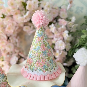 May include: A set of three pink and white party hats with a floral print and a pink pom-pom on top. The hats are sitting on a white cake stand.