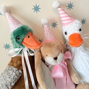 Puede incluir: Tres juguetes de peluche: un pato, un conejito y un ganso, cada uno con un gorro de fiesta. El pato tiene cabeza verde y pico naranja, el conejito tiene orejas largas y el ganso tiene pico naranja. Todos tienen cintas y lazos blancos.