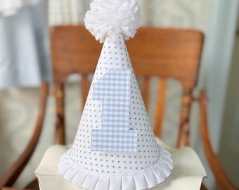 May include: A white and blue party hat with a white pom-pom on top. The hat has a blue gingham number "1" on the front and blue polka dots on the rest of the hat.