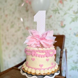 May include: A single-tiered white cake with pink frosting and a pink bow on top. The cake is decorated with pink hearts and a pink number one topper.