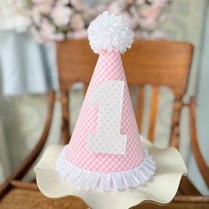 Pode incluir: Chapéu de aniversário em vichy rosa e branco com um pompom branco no topo e um número "1" de feltro branco com bolinhas rosa.