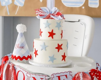Può includere: Una torta bianca a due piani decorata con stelle rosse e blu, sormontata da un numero uno e un fiocco decorativo. Sono presenti anche uno striscione con la scritta "ONE" e un cappello da festa, il tutto a tema patriottico.