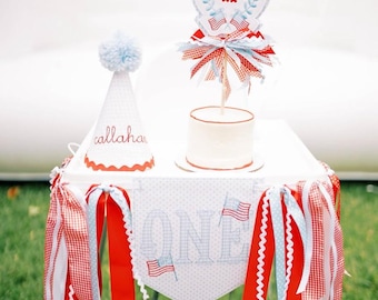 Può includere: Un tavolo bianco decorato per una festa di primo compleanno. Presenta una torta, un cappello da festa con il nome "Callahan" e uno striscione con la scritta "ONE" con bandiere americane. Nastri rossi, bianchi e blu adornano il tavolo.
