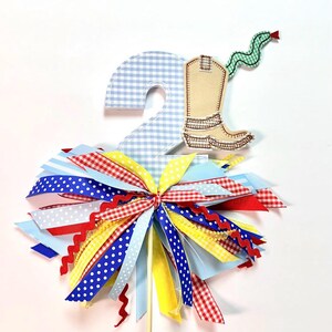 Puede incluir: Adorno festivo para tarta con un número 2 a cuadros, una bota de vaquero y una serpiente verde. La base es una colorida variedad de cintas rojas, blancas, azules y amarillas, ideal para una fiesta de cumpleaños.