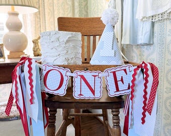 Peut inclure: Une chaise haute en bois décorée pour un premier anniversaire. Une bannière épelle "ONE" en lettres rouges sur un fond à carreaux bleu. Un petit gâteau blanc et un chapeau de fête avec un pompon sont posés sur la chaise. Des rubans rouges et bleus pendent de la chaise.