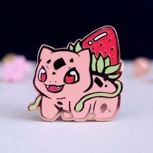 May include: A rose gold enamel pin featuring a cartoon pink Pokemon with a red strawberry on its back. The Pokemon has green leaves and a tail.