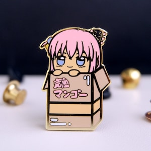 May include: A gold-colored enamel pin featuring a cartoon character with pink hair, blue eyes, and a pink bow. The character is peeking out of a brown cardboard box with the Japanese text "激熱 マツコー" printed on the front.