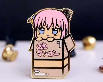 Puede incluir: Un pin de esmalte dorado con un personaje de dibujos animados con cabello rosado, ojos azules y un lazo rosa. El personaje asoma desde una caja de cartón marrón con el texto japonés "激熱 マツコー" impreso en la parte delantera.