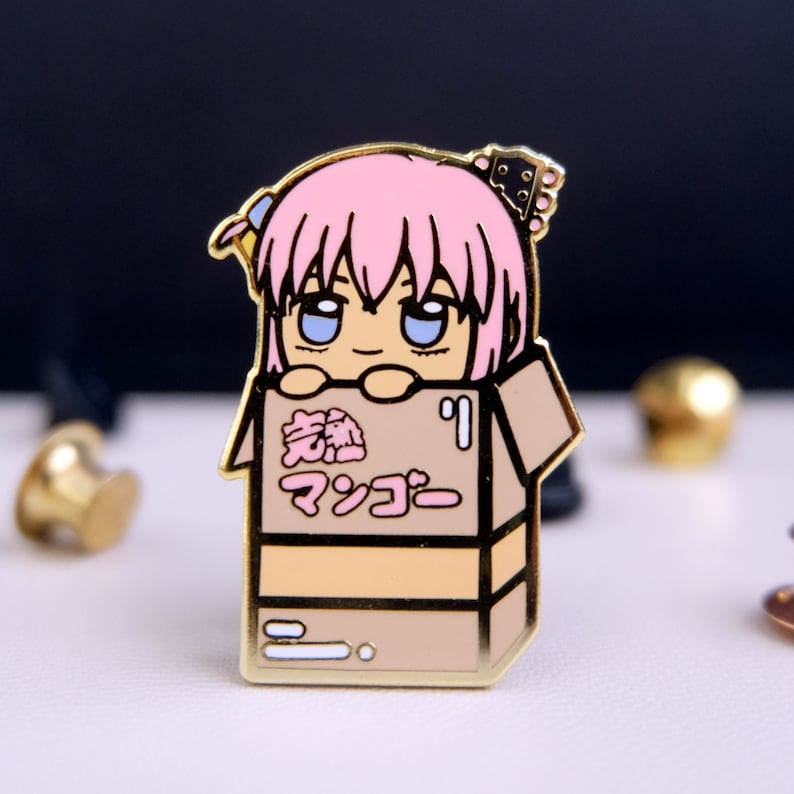 Puede incluir: Un pin de esmalte dorado con un personaje de dibujos animados con cabello rosado, ojos azules y un lazo rosa. El personaje asoma desde una caja de cart&oacute;n marr&oacute;n con el texto japon&eacute;s "激熱 マツコー" impreso en la parte delantera.