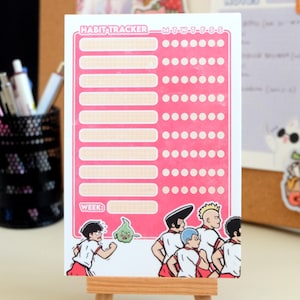May include: A pink and white habit tracker with a grid of circles for each day of the week. The tracker has a section for writing in habits and a section for writing in the week number. The tracker has a cartoon illustration of a group of people running with a green ghost.