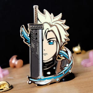 Peut inclure: Une broche en émail métallique présentant un design divisé avec un personnage aux cheveux blonds en pointe et un œil bleu d'un côté, et une grande épée gris foncé aux détails mécaniques de l'autre, ornée de motifs d'énergie bleus et blancs.