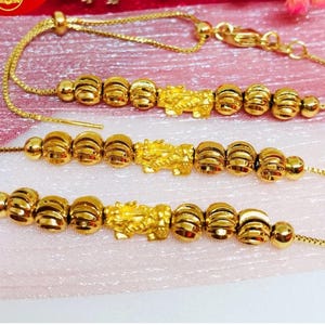 Puede incluir: Tres pulseras de oro con un diseño de cuentas y un pequeño dije de dragón de oro en cada una. Las pulseras están sobre un fondo rosa y blanco.