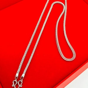Puede incluir: Un collar plateado con un diseño de cadena trenzada y un cierre de mosquetón. El collar se muestra sobre una superficie roja, creando un contraste que resalta los detalles de la joya. La cadena está hecha de pequeños eslabones interconectados.