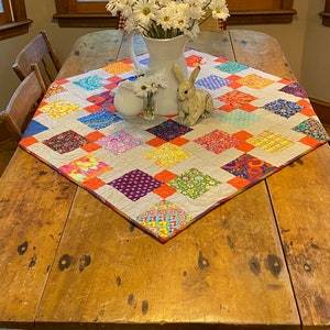 May include: A colorful patchwork quilt table runner with a white background and orange, red, purple, blue, and green squares. A white pitcher with a bouquet of white daisies and a white ceramic bunny figurine are on top of the runner.