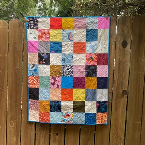 May include: A colorful patchwork quilt with a variety of patterns and colors, including pink, blue, yellow, orange, and green. The quilt is hanging on a wooden fence.