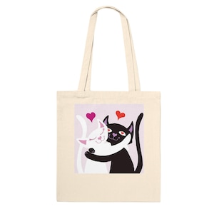 May include: A cream-coloured tote bag with a graphic of two hugging cats, one white and one black, with pink and red hearts above them. The cats are smiling, set against a light purple background.