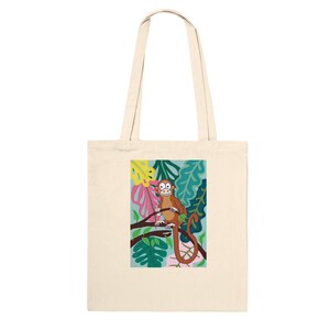 May include: A cream-coloured tote bag with a vibrant illustration of a monkey on a branch. The monkey is brown, with a long tail, set against a backdrop of green, blue, pink, and yellow leaves. The bag has two cream-coloured handles.
