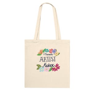 May include: Cream-coloured tote bag featuring the text "Planner Artist Maker" in a playful font, decorated with colourful floral accents. The bag has two long handles and is made of a durable fabric.