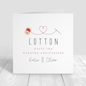 May include: White anniversary card featuring a red cotton reel, a heart, and a needle illustration. The word "COTTON" is above "HAPPY 2ND WEDDING ANNIVERSARY" and the names "Katie & Oliver".