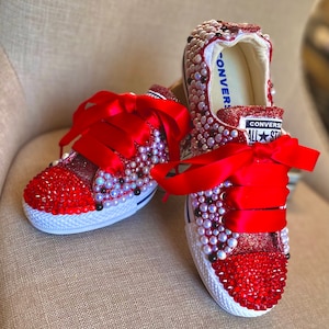 Könnte beinhalten: Rote Glitzer-Converse-Sneaker mit roten Schleifenbändern und roten und weißen Strasssteinen verziert. Die Sneaker haben den Text "CONVERSE" und "ALL STAR" aufgedruckt.