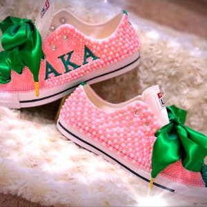 Puede incluir: Zapatillas rosas y verdes con un lazo verde y las letras "AKA" en verde en el lateral. Las zapatillas están cubiertas de perlas rosas.