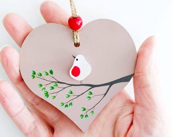 May include: A heart-shaped wooden decoration featuring a painted bird on a branch. The bird is white with a red breast. The ornament is taupe with green leaves and a brown branch. A red bead and jute string are attached.