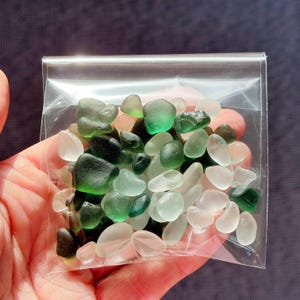 May include: A clear plastic bag filled with sea glass in shades of green, white, and black. The sea glass pieces are smooth and rounded, with varying sizes and shapes. The bag is held in a person's hand.
